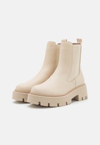 Bottines beige avec une finition en cuir lisse, des panneaux latéraux élastiques et une semelle compensée texturée avec des découpes.