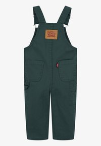 Tmavě zelené bavlněné kalhoty s laclem, vybavené nastavitelnými ramínky, čtvercovou kapsou s značkovou nášivkou a dvěma bočními kapsami.