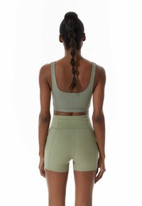 Mujer de piel oscura y con largas trenzas de cabello se encuentra de espaldas, vistiendo un sujetador deportivo verde claro y unos pantalones cortos de talle alto a juego.