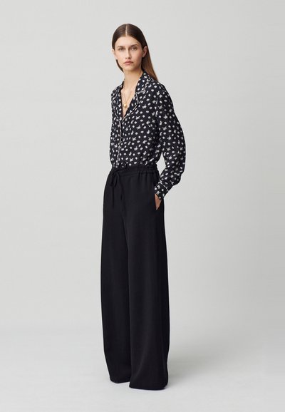 Blusa nera a maniche lunghe con motivi floreali bianchi, abbinata a pantaloni neri a gamba larga con vita alta, dotati di cinturino elastico e tasche.