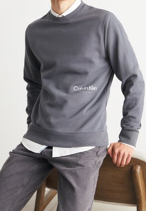 Mand iført grå Calvin Klein sweatshirt over hvid skjorte, siddende på en træstol med den ene hånd hvilende på ryglænet.