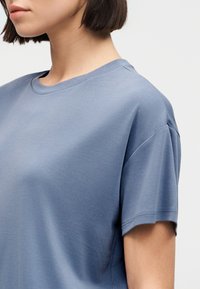 Γυναίκα με κοντά καστανά μαλλιά που φοράει απλό ανοιχτό μπλε κοντομάνικο μπλουζάκι με στρογγυλή λαιμόκοψη, φαίνεται από το στήθος και πάνω σε λευκό φόντο.