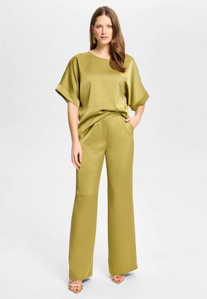 Vrouw staand in olijfgroene satijnen wijde broek en bijpassende blouse met korte mouwen, met lang haar en gouden oorbellen, tegen een witte achtergrond.