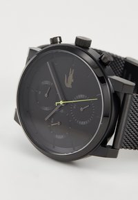 Montre chronographe noire avec un bracelet en maille métallique texturé, boîtier rond et trois sous-cadrans. Caractéristiques d'une aiguille des secondes verte et d'un logo argenté.