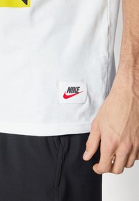 White cotton t-shirt featuring a yellow graphic and black lettering on the front, with a red and black Nike logo patch on the hem.