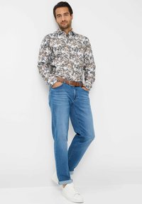 Camicia a maniche lunghe con motivo floreale in colori tenui, abbinata a jeans in denim blu chiaro e sneakers bianche. La cintura marrone aggiunge un contrasto.