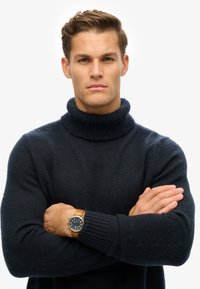 Suéter de cuello alto en azul marino oscuro, con canalé en el cuello y los puños. Llevado con un reloj dorado y azul marino, con los brazos cruzados. Tejido simple y texturizado.