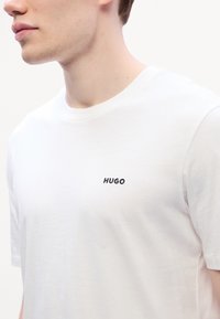 Hvit bomulls t-skjorte med rund hals og korte ermer, med "HUGO" trykket i svart på venstre brystområde.