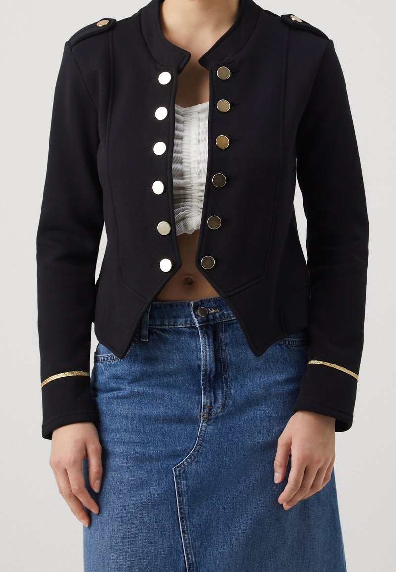 Femme portant une veste militaire noire avec des boutons et des bordures dorés, par-dessus un crop top blanc froncé et une jupe en denim bleu.