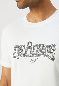 Camiseta de algodón blanca con un logo "NIKE" delineado en negro impreso en el pecho, con un pequeño swoosh de Nike debajo.