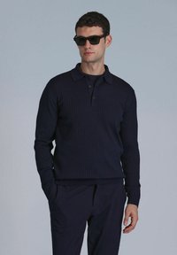 Marineblaues geripptes Strickpoloshirt mit Kragen und drei Knöpfen. Kombiniert mit passenden marineblauen Hosen, dazu dunkle Sonnenbrille.
