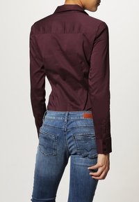 Camisa de manga larga color burdeos con un diseño ajustado, combinada con unos vaqueros ajustados azules que presentan una textura desgastada y detalles de costura en los bolsillos traseros.