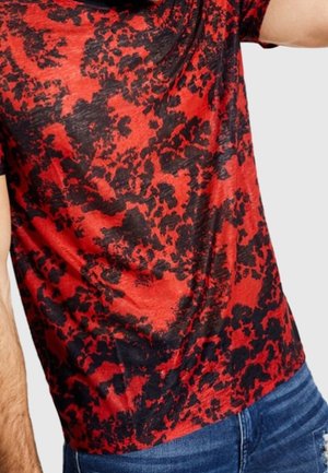 Chemise à manches courtes à motif abstrait rouge et noir portée avec un jean bleu, montrée de la moitié du torse jusqu’au haut des cuisses.