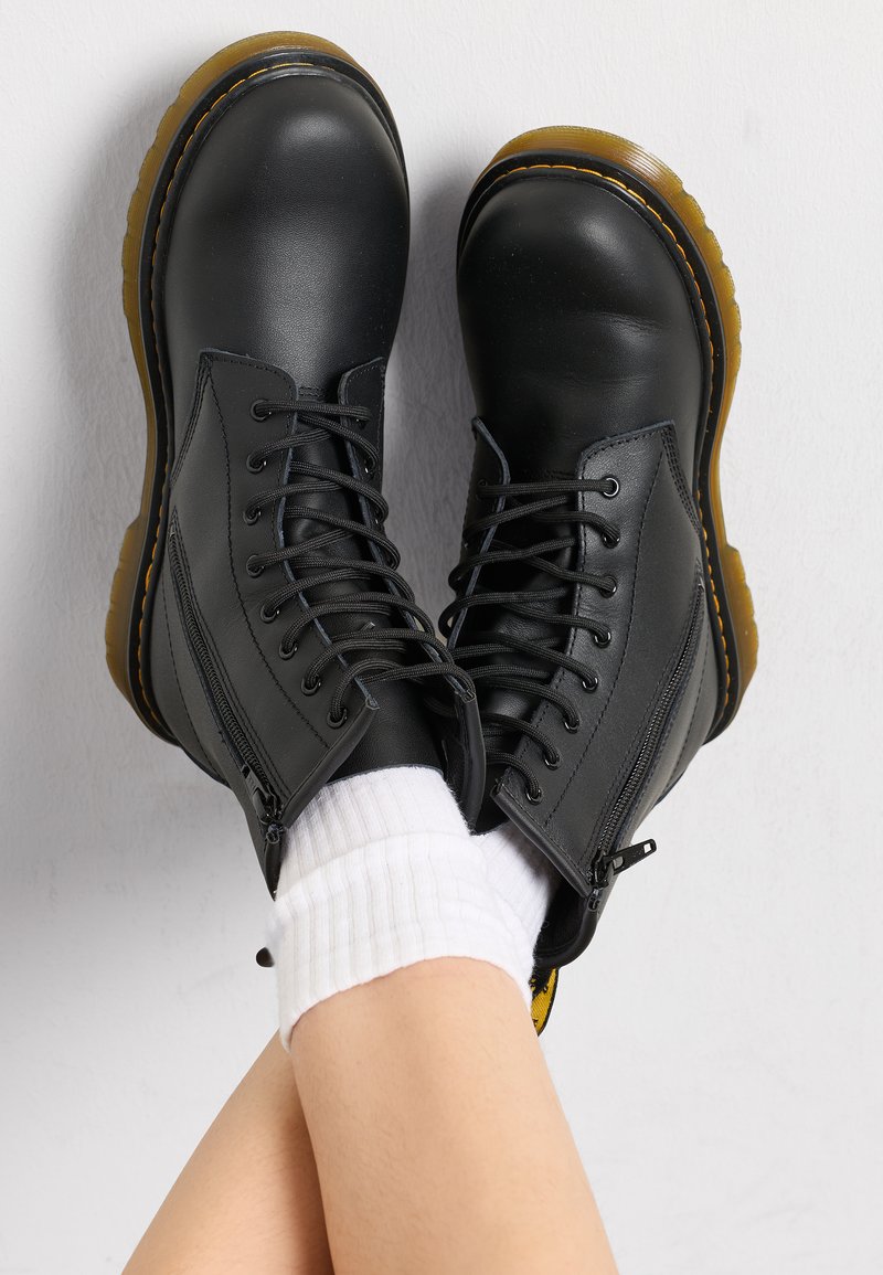 Botas de couro pretas com biqueira redonda, frente com atacadores, fecho com zíper e sola amarela em contraste. Superfície texturizada, design robusto.
