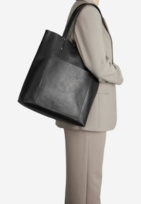 Juodas odinis tote krepšys su lygiu paviršiumi, ilgomis rankenomis ir didele priekiu kišene. Paprasta stačiakampio formos konstrukcija, be matomų detalių ar puošmenų.