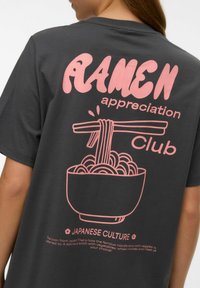 Węglowoszara koszulka z dużym różowym nadrukiem miski ramenu z pałeczkami, nad którą znajduje się napis "Klub Miłośników Ramenu".