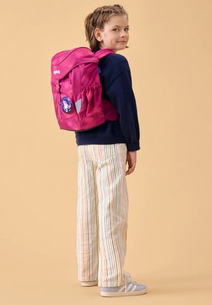Junges Mädchen mit geflochtenem Haar, das einen marineblauen Pullover, gestreifte Hosen, Turnschuhe und einen leuchtend pinken Rucksack mit Einhorn-Aufnäher trägt, seitlich stehend.