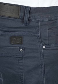 Donkerblauwe spijkerbroek met een gestructureerde stof, voorzien van een leren label, meerdere zakken en zilverkleurige metalen accenten.