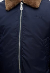 Veste bleu marine avec col en fourrure marron, fermeture éclair argentée au centre et logo Schott NYC brodé sur la poitrine.