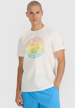 Mężczyzna z krótkimi włosami, ubrany w kremowy t-shirt z grafiką palmy i napisem „Free on Board Chasing Waves”, w połączeniu z jasnoniebieskimi szortami.
