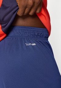 Χέρι με σκούρο δέρμα σηκώνει κόκκινο και μπλε πουκάμισο, αποκαλύπτοντας τη μέση και μπλε ελαστικά σορτς με λευκό λογότυπο Climacool.