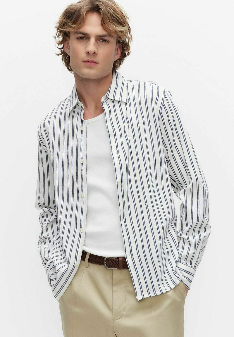 Jonge man met golvend blond haar, draagt een open wit overhemd met marineblauwe strepen, een wit hemdje, beige broek en een bruine gevlochten riem.