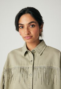 Kaki spalva striukė su šoniniu dizainu, papuoštu sidabro grandinėlėmis. Ji turi mygtukų užsegimą ir klasikinį apykaklę. Lygus medvilnės audinys.