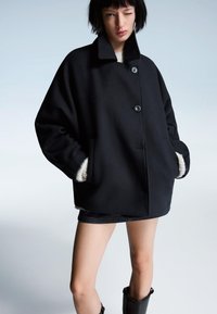 Črn prevelik plašč z visokim ovratnikom, dvema sprednjima žepoma in kremnim podloženjem; ima dva velika gumba spredaj.