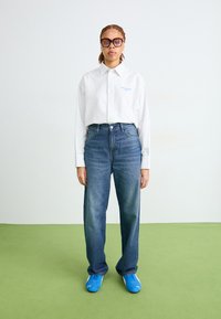 Camisa blanca a rayas con botones, vaqueros de tiro alto azules y zapatillas brillantes azules, de pie sobre un suelo verde contra un fondo liso.