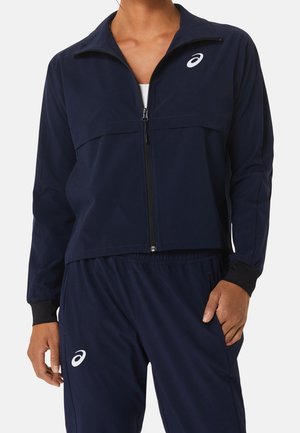 Person trägt eine marineblaue Asics Zip-up-Jacke und passende Hose, steht mit einer Hand in der Tasche vor weißem Hintergrund.