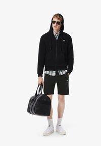 Zwarte zip-up hoodie, geruit overhemd, donkergroene shorts, witte sneakers en een grijze tas. Model draagt een zonnebril; heeft Lacoste-logo's.