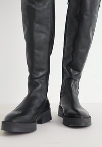 Bottes en cuir noir au-dessus du genou avec une texture lisse, une semelle plate robuste et des détails de fermeture éclair sur le côté. Design simple et épuré.