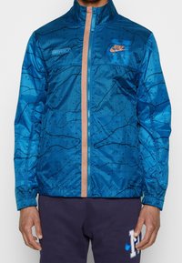 Sininen takki, jossa on geometrinen kuvio, vetoketjusuljenta ja kontrastivärinen pinkki yksityiskohta. Kevyt materiaali ja joustavat hihansuut.