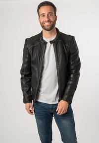 Giacca in pelle nera con accenti texturizzati sulle spalle, slim fit e chiusura con zip. Indossata sopra una camicia bianca e abbinata a jeans blu.