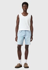 Canottiera bianca in maglia con texture a righe verticali, pantaloni corti in denim blu chiaro, scarpe slip-on marroni e calzini bianchi alla caviglia.