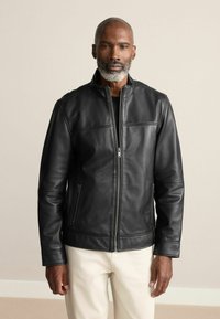 Schwarze Lederjacke mit Stehkragen, Reißverschluss und zwei seitlichen Taschen. Glatte Textur und dezente Nähdetails.