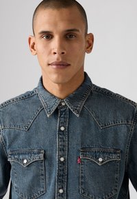 Cămașă din denim albastră, cu închidere prin capse, două buzunare pe piept, yoke în stil Western și accente de cusătură albă.