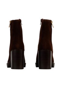 Botines de ante marrón con un acabado texturizado, tacones cuadrados y detalles de cremallera dorada en la parte trasera. Diseño minimalista y de color sólido.