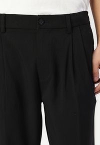 Pantaloni neri in tessuto testurizzato, con chiusura a bottone, pieghe frontali e tasche laterali. La vita è piatta e semplice.