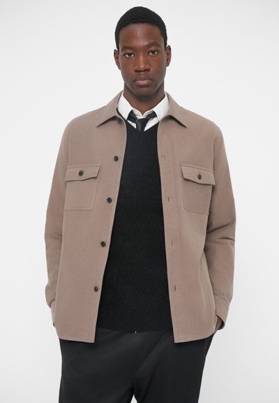 Brun, knappingsjakke med to brystlommer og spiss krage, lagd over en svart genser og hvit skjorte. Glatt tekstur, skreddersydd passform.