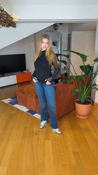 Black polka dot top with sheer sleeves, blue flared jeans, and silver sneakers. Indoor setting with a green plant and orange couch.