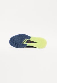 Semelle de chaussure de tennis en noir et jaune néon avec un motif en zigzag. Présente une traction en caoutchouc bleu sur le dessous et le logo de la marque 'HEAD' visible.