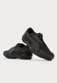 Paire de chaussures de sport noires avec semelles texturées et une bride auto-agrippante, positionnée sur un fond blanc.
