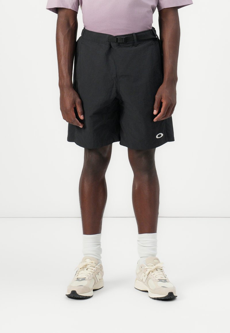 Schwarze Shorts aus leichtem Stoff. Sie verfügen über einen Gürtel, Taschen an den Seiten und ein kleines kreisförmiges Logo am Saum. Kombiniert mit weißen Socken und Turnschuhen.