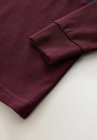 Burgunderfarvet langærmet skjorte med ribbede manchetter og struktureret stof. Sømmen er lige, og materialet har et blødt, vævet udseende.