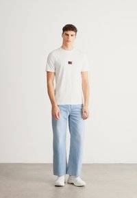 Λευκό μπλουζάκι με κοντό μανίκι και μικρό κόκκινο και μαύρο σήμα στο στήθος, συνδυασμένο με ανοιχτό μπλε τζιν παντελόνι με φαρδιά μπατζάκια και λευκά αθλητικά παπούτσια.