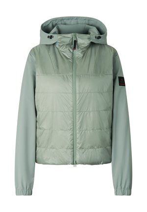 Junge Frau trägt grüne gesteppte Kapuzenjacke, passende grüne Leggings mit schwarzen Einsätzen und schwarze Wanderschuhe, steht frontal.