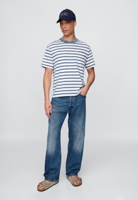 Stripete kortermet t-skjorte i hvitt og marineblått, kombinert med lyseblå jeans og grå sandaler med stropp, toppet med en marineblå caps.