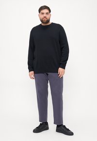 Pulover negru cu guler rotund, asociat cu pantaloni de culoare mov deschis. Modelul poartă adidași negri; fundalul este alb simplu.