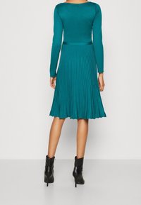 Robe longue teal à manches longues avec un haut ajusté et une jupe plissée, dotée d'une taille cintrée et de bottes noires à talons hauts jusqu'aux chevilles. Texture lisse.